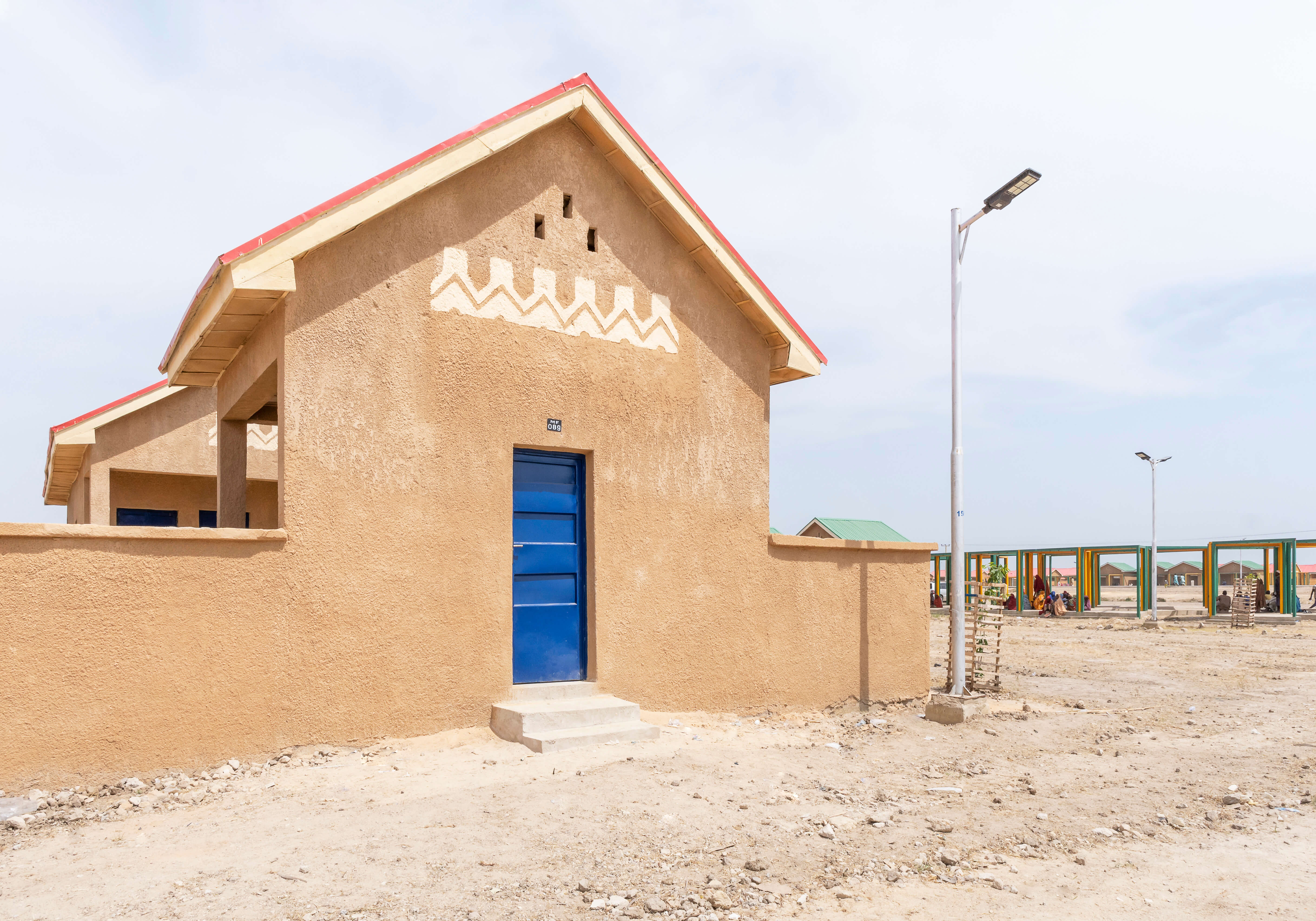 A housing project in Ngarannam, showcasing the design by Oshinowo Studio, a leading architectural design studio in Lagos, led by Tosin Oshinowo. The image features a row of new homes with sandy ochre finishes and distinct red roofs. The façades are decorated with traditional white geometric patterns above vibrant blue doors and matching window shutters, standing out against the clear sky. The structures are modern yet reflect local architectural styles, set within a dry landscape.