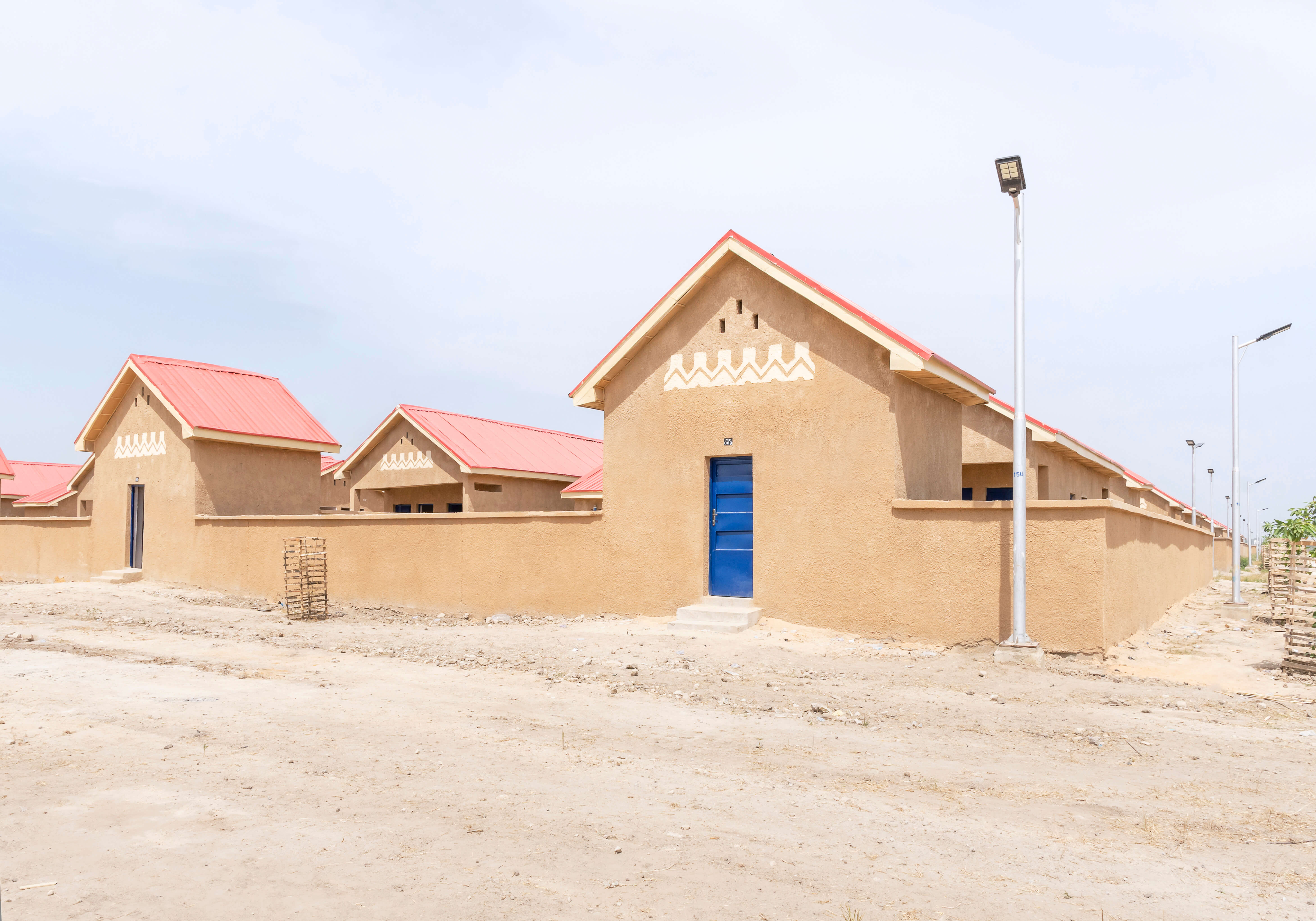 Architectural photograph of a housing project in Ngarannam, showcasing the design by Oshinowo Studio, a leading architectural design studio in Lagos, led by Tosin Oshinowo. The image features a row of new homes with sandy ochre finishes and distinct red roofs. The façades are decorated with traditional white geometric patterns above vibrant blue doors and matching window shutters, standing out against the clear sky. The structures are modern yet reflect local architectural styles, set within a dry landscape.