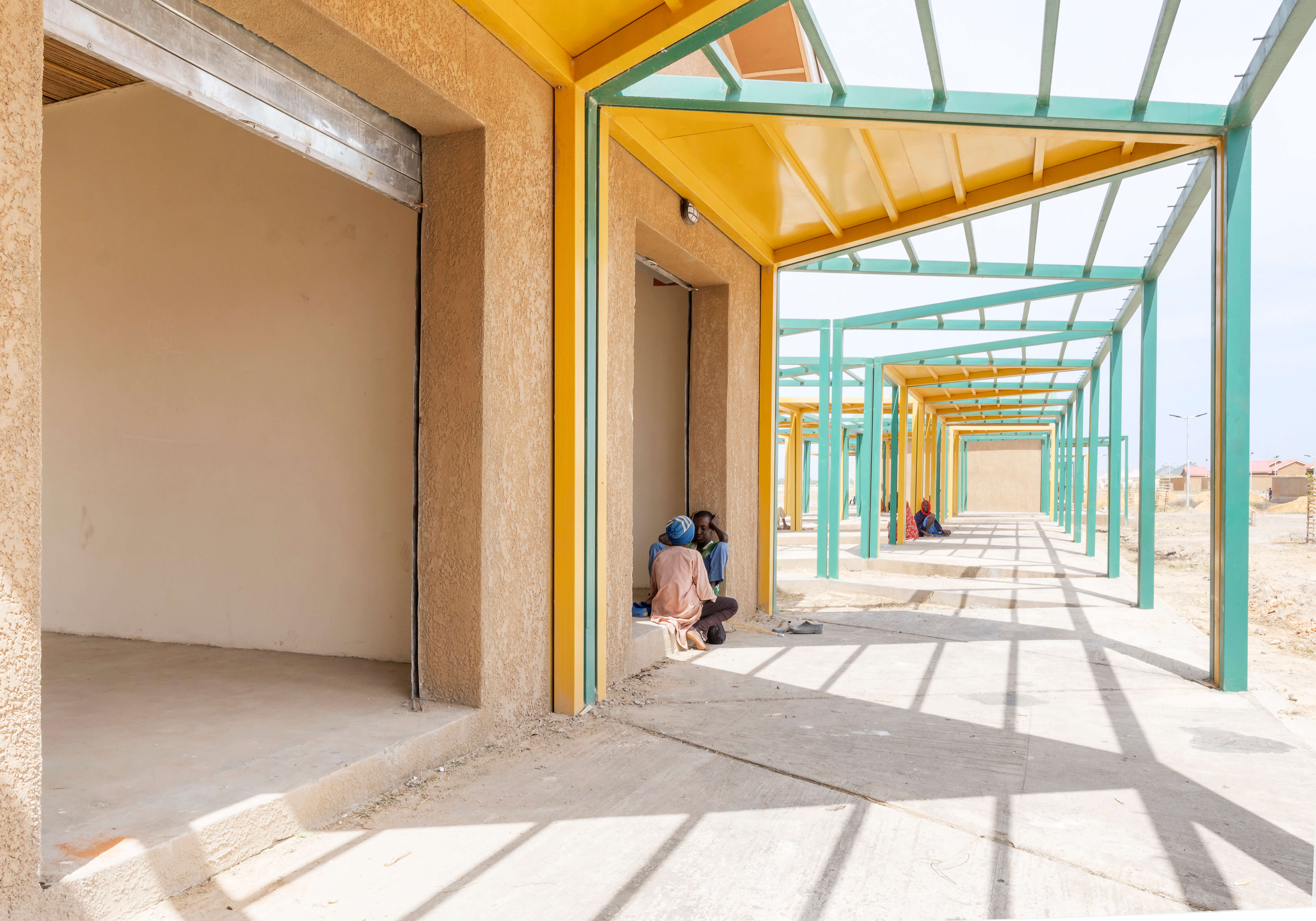 Image showing a sunny, open-air corridor of a market complex designed by Oshinowo Studio, a leading architectural design studio in Lagos, led by Tosin Oshinowo. The corridor features a series of bold green and yellow metal columns and beams that create a linear perspective, leading the eye through the space. Locals sit in the shade provided by the structure, against the backdrop of shops with ochre walls and thatched roofs. The space is filled with natural light and shadows, highlighting the geometric beauty of the architecture.