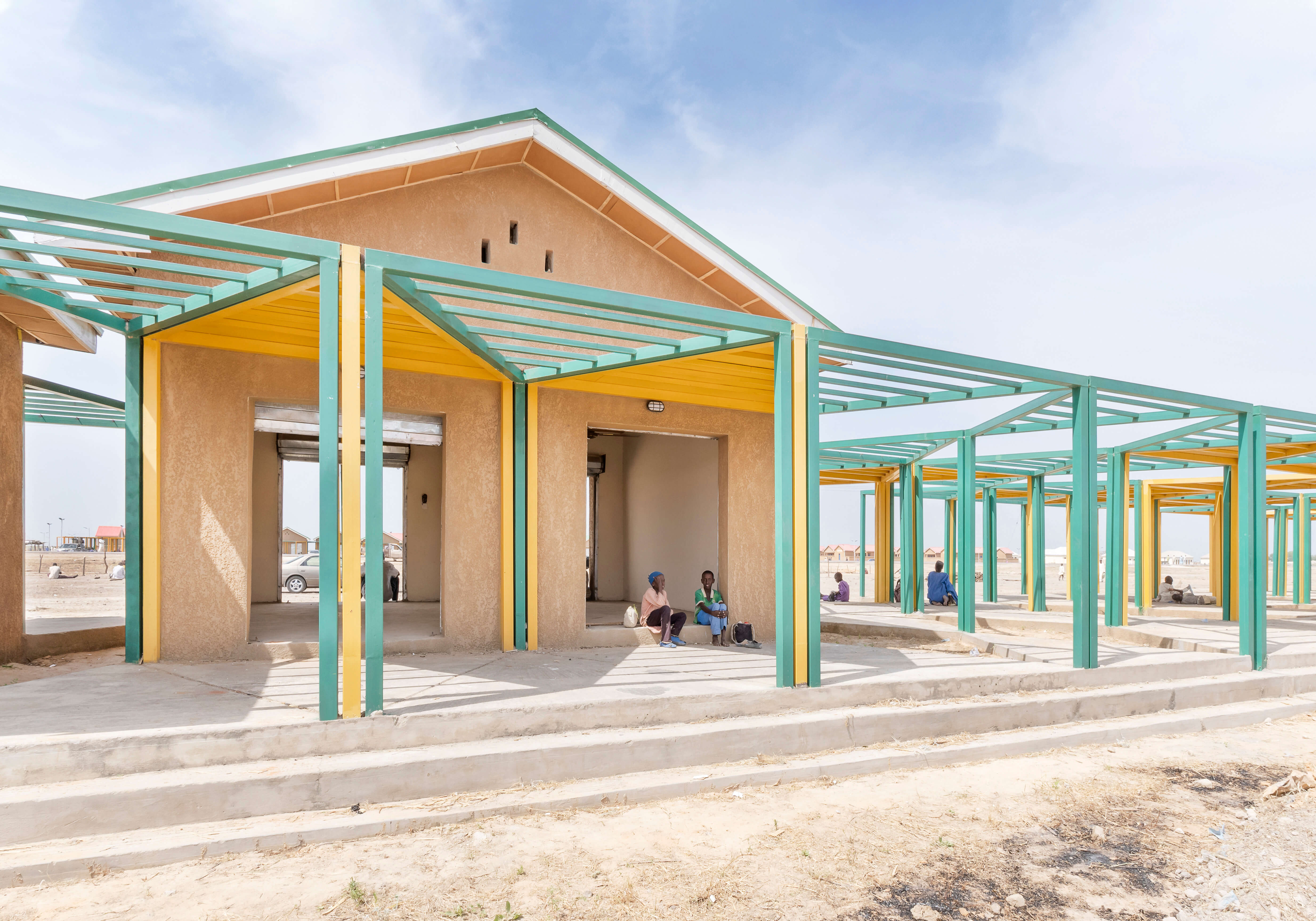 A spacious, well-ventilated marketplace shelter in a sandy environment, designed by Oshinowo Studio, a leading architectural design studio in Lagos, led by Tosin Oshinowo. The structure is characterized by a sequence of bright yellow and green steel beams supporting the roof, harmonizing with the natural-colored walls. Locals gather under the protection of the canopy, enjoying the communal space, with the clear sky above hinting at the open-air design.