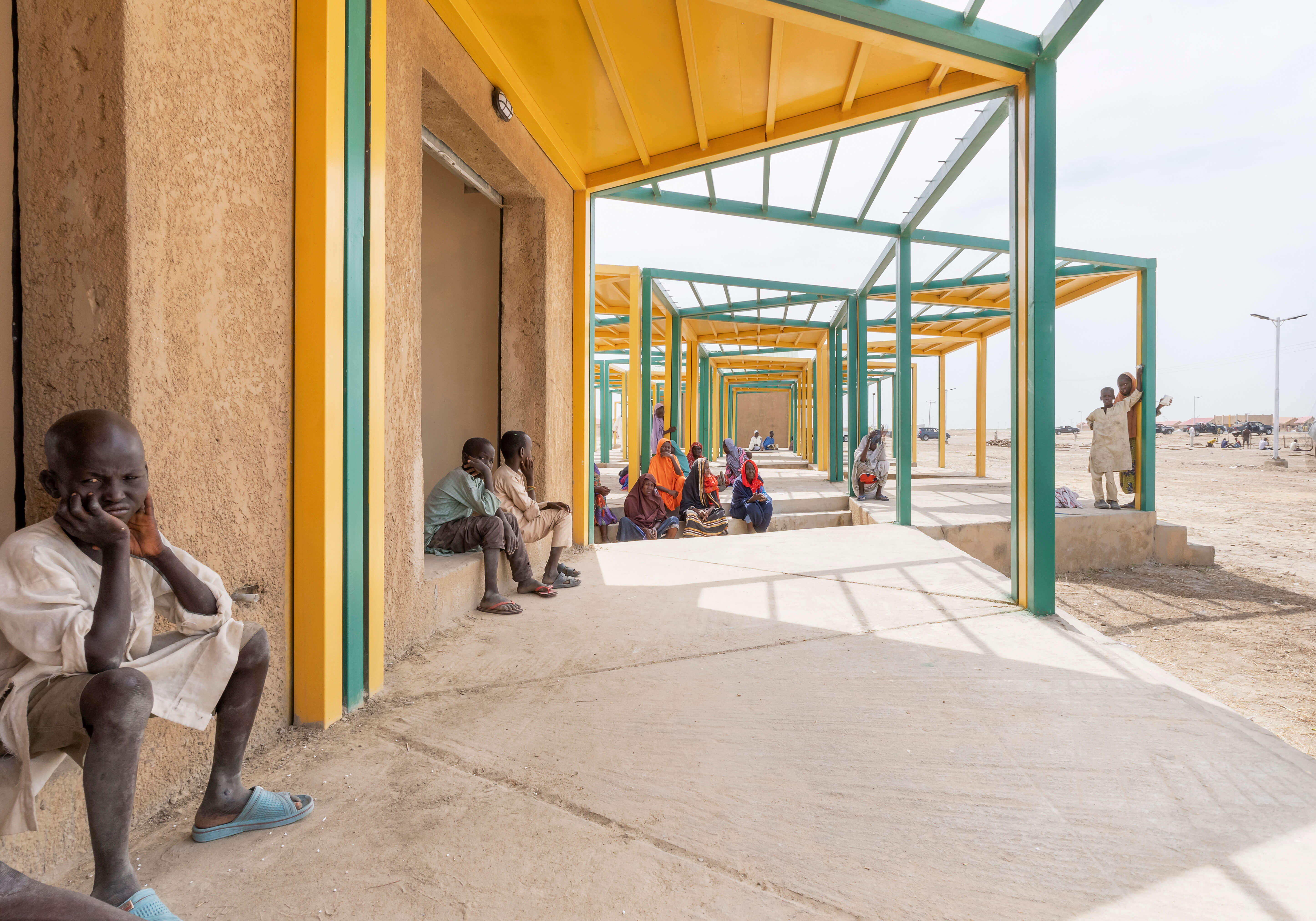 Image of a communal shelter designed by Oshinowo Studio, a leading architectural design studio in Lagos, led by Tosin Oshinowo, providing a shaded gathering space for the local community. The structure is composed of a series of turquoise and yellow pillars that support the roof, creating a long corridor. Individuals are seated along the walls, engaging in social activities, while a child playfully interacts with the camera. The design offers a welcoming environment that facilitates community interactions and daily life.