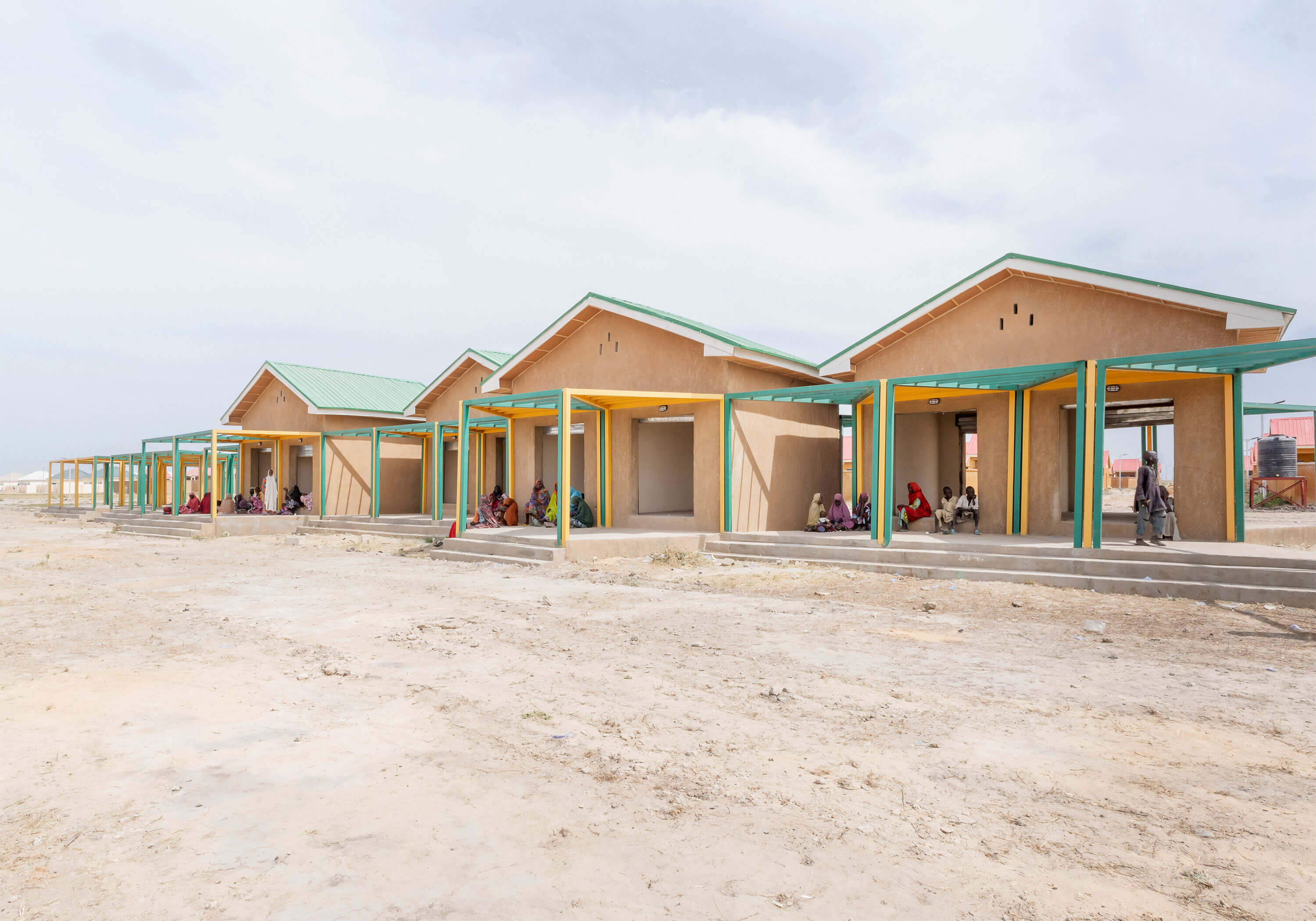A view of a community marketplace in a sandy, open area, featuring design elements from Oshinowo Studio, a leading architectural design studio in Lagos, led by Tosin Oshinowo. The market includes several ochre-colored buildings with green metal roofs, complemented by yellow and green pillars. Local people are gathered in small groups, seated on benches and ledges under the shade of the overhanging roofs, while children play in the foreground, animating the serene communal space.