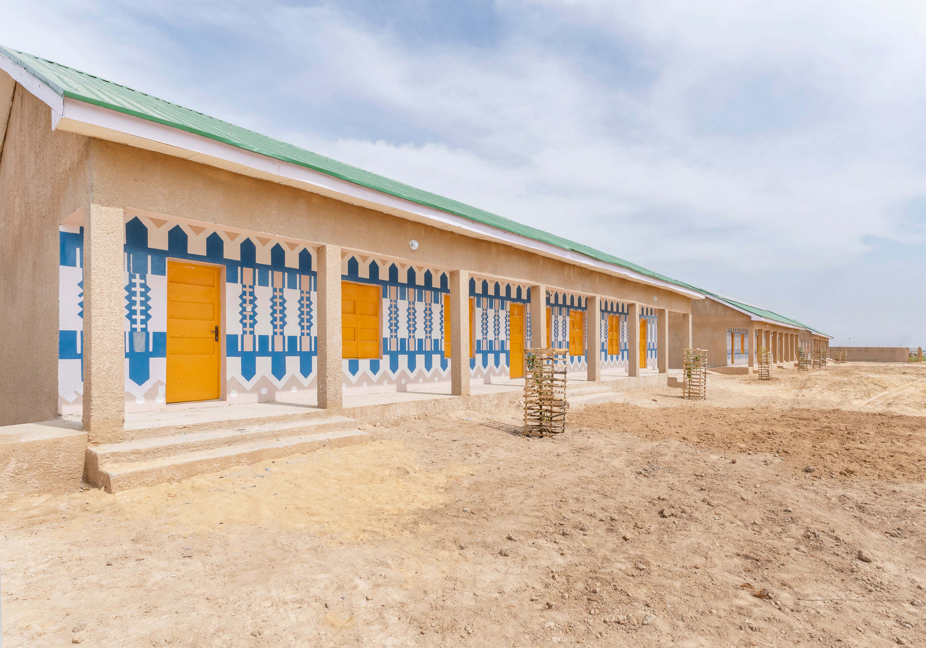 A long building in a desert landscape, showcasing the vibrant and culturally-inspired architectural style of Oshinowo Studio, a leading architectural design studio in Lagos, led by Tosin Oshinowo. The facade is decorated with blue and white geometric patterns surrounding yellow doors and ornate window shutters, set against ochre walls and under a green metal roof. The building, possibly a school or community center, brings a burst of color and design to the sandy surroundings.