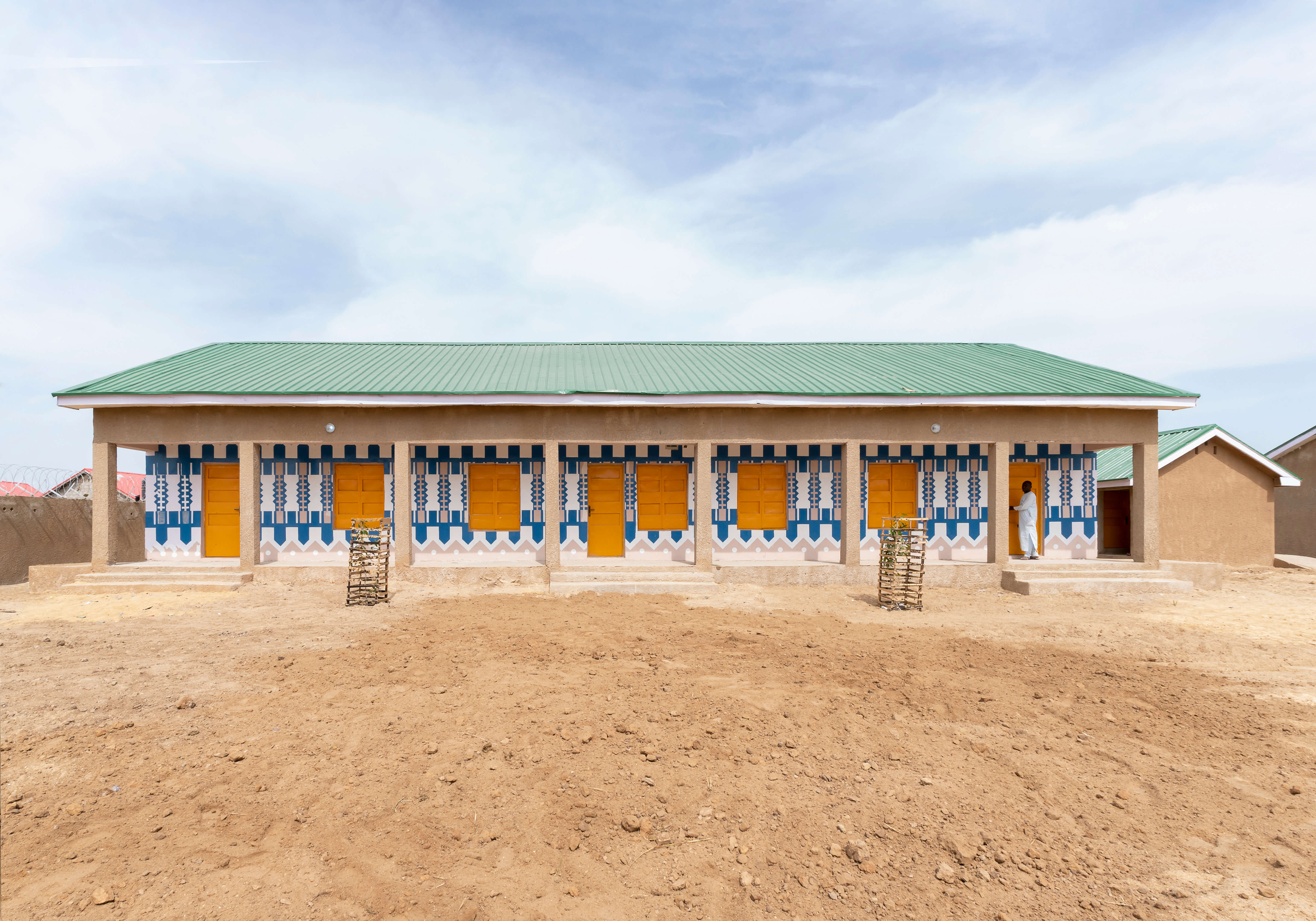 A long building in a desert landscape, showcasing the vibrant and culturally-inspired architectural style of Oshinowo Studio, a leading architectural design studio in Lagos, led by Tosin Oshinowo. The facade is decorated with blue and white geometric patterns surrounding yellow doors and ornate window shutters, set against ochre walls and under a green metal roof. The building, possibly a school or community center, a person standing in front of door, brings a burst of color and design to the sandy surroundings.
