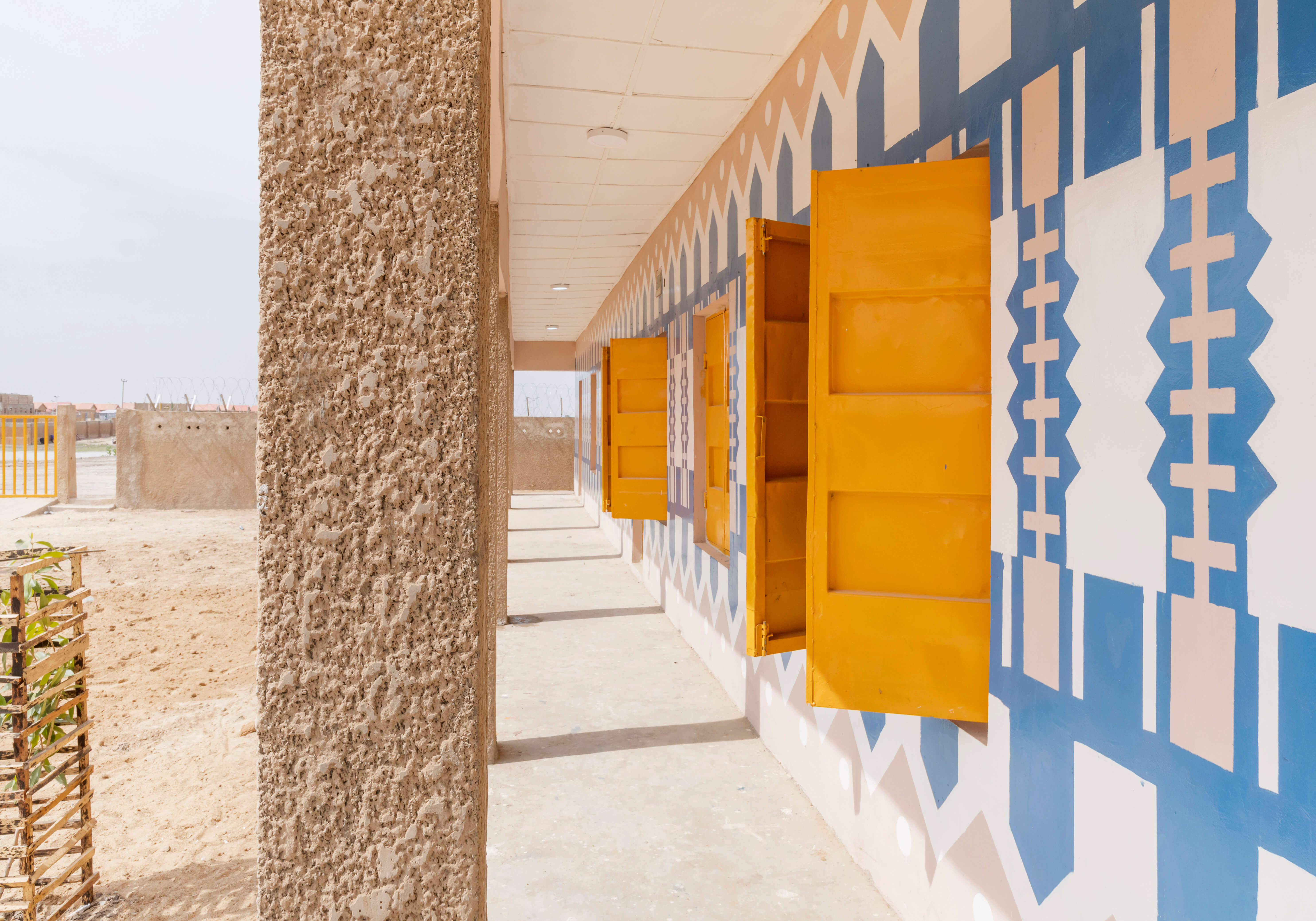 The image captures a part of a vibrant marketplace designed by Oshinowo Studio, a leading architectural design studio in Lagos, led by Tosin Oshinowo, characterized by a bright color scheme and bold geometric patterns. The marketplace features a sandy pathway leading to a series of shops with blue and white patterned walls and vivid yellow doors, reflecting a modern take on traditional African aesthetics. The space is open and airy, promoting a welcoming atmosphere for community gathering and commerce.