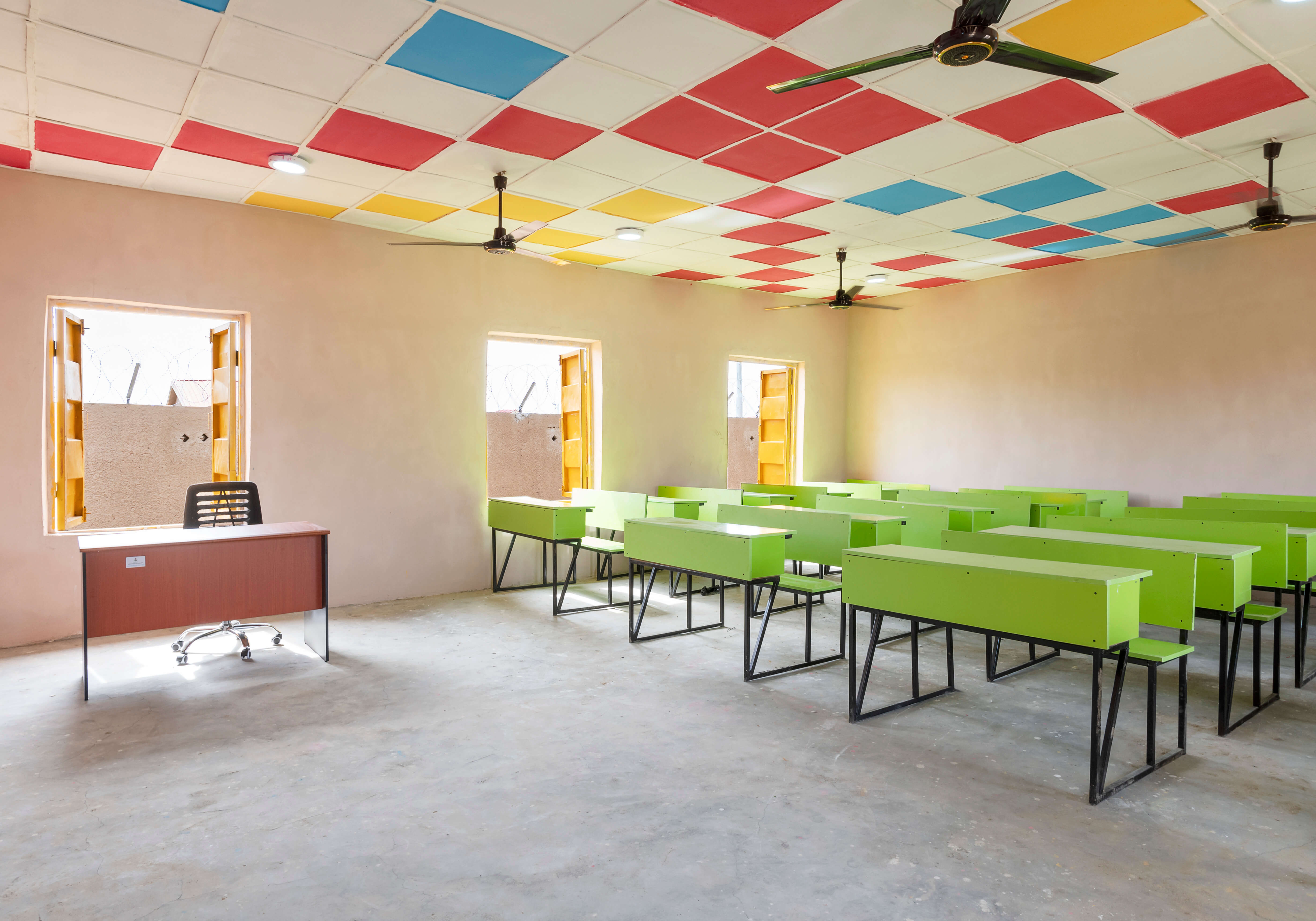 Interior of a brightly colored classroom designed by Oshinowo Studio, a leading architectural design studio in Lagos, led by Tosin Oshinowo, displaying a playful and inviting learning environment. The ceiling is adorned with a patchwork of red, blue, and yellow tiles, while the room is furnished with vibrant green tables and black chairs. Ample natural light streams in through the open windows, illuminating the space and the solitary desk at the front.