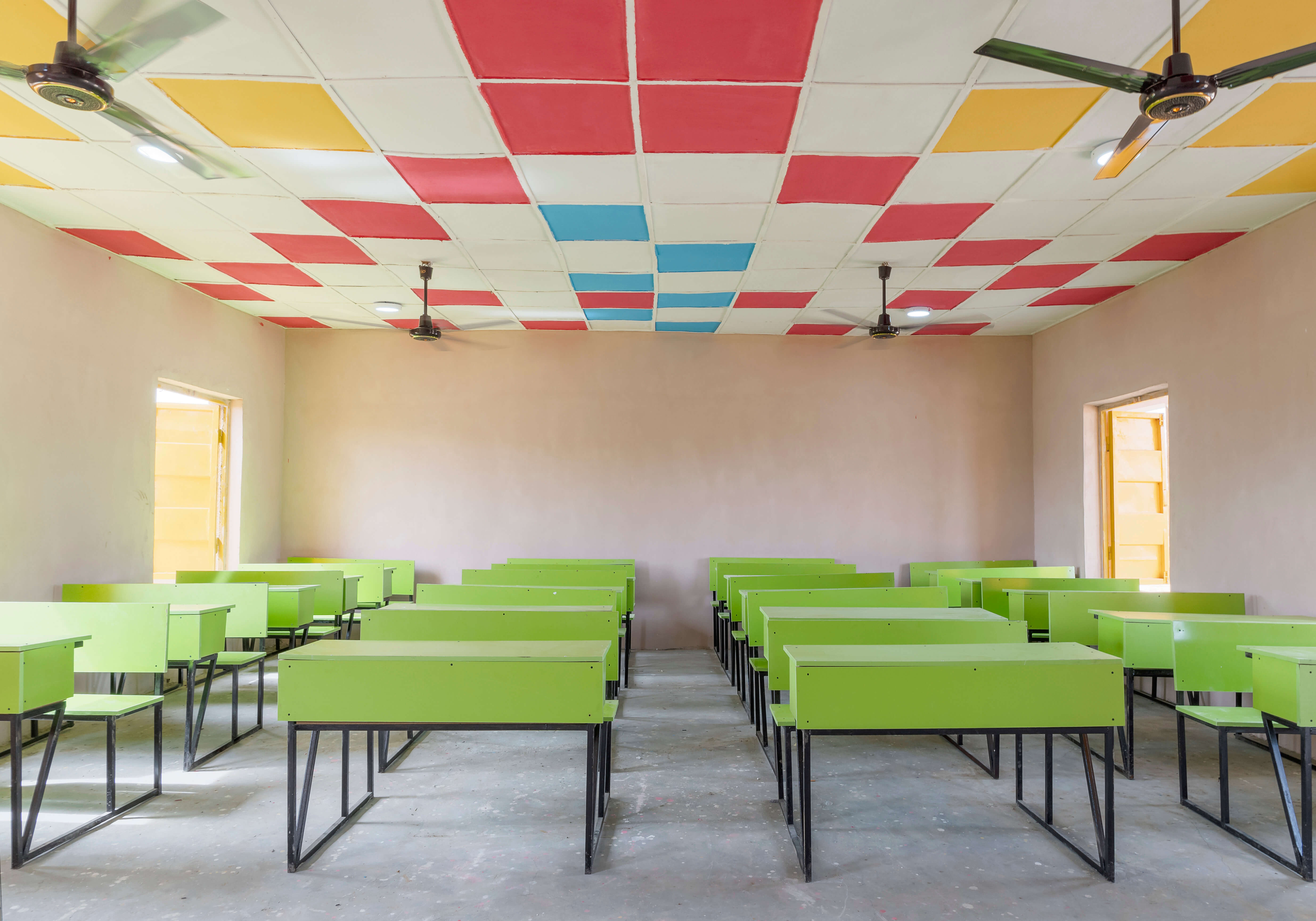 Front view of the interior of a brightly colored classroom designed by Oshinowo Studio, a leading architectural design studio in Lagos, led by Tosin Oshinowo, displaying a playful and inviting learning environment. The ceiling is adorned with a patchwork of red, blue, and yellow tiles, while the room is furnished with vibrant green tables and black chairs. Ample natural light streams in through the open windows, illuminating the space.