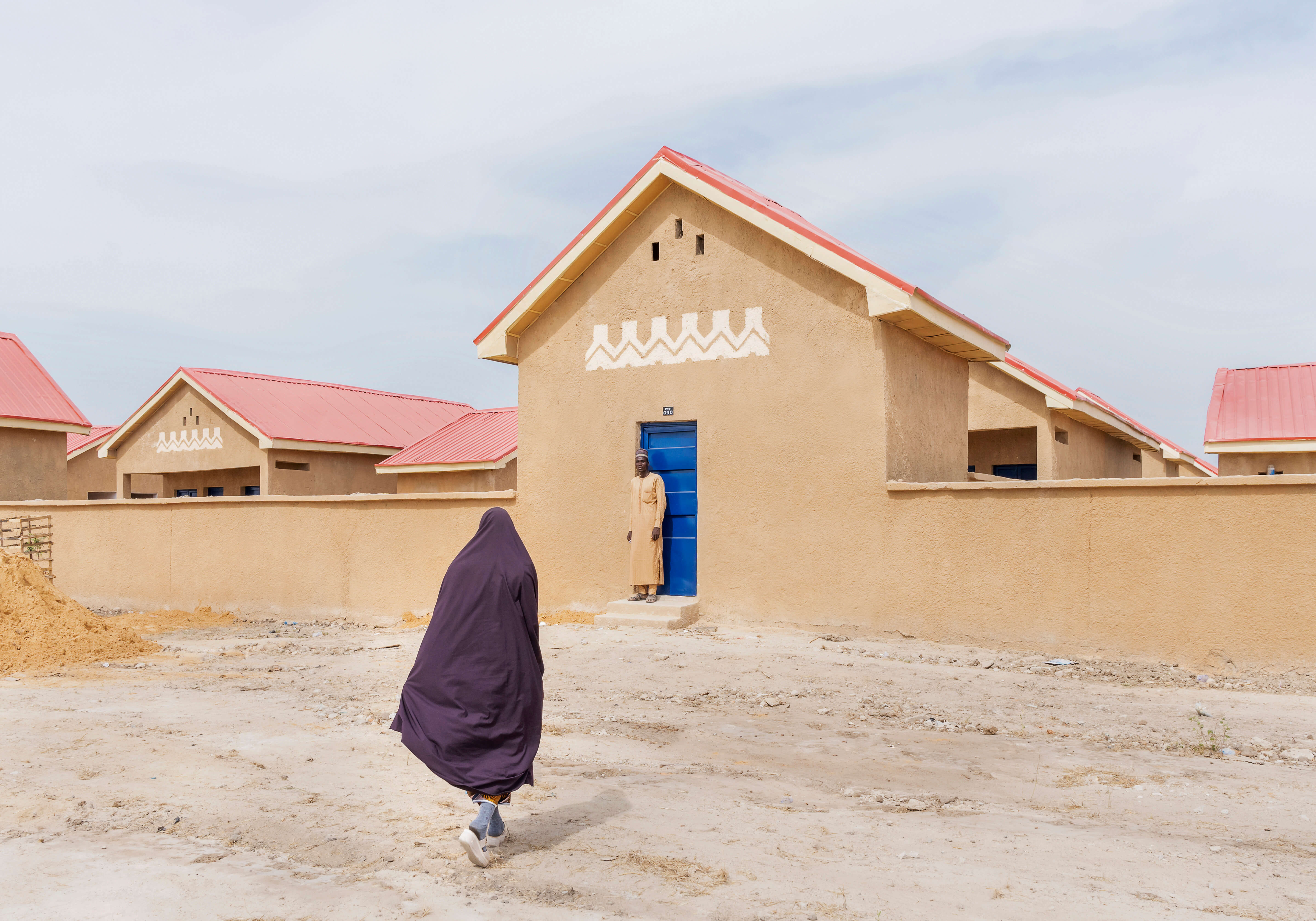 A housing project in Ngarannam, showcasing the design by Oshinowo Studio, a leading architectural design studio in Lagos, led by Tosin Oshinowo. The image features a row of new homes with sandy ochre finishes and distinct red roofs. The façades are decorated with traditional white geometric patterns above vibrant blue doors and matching window shutters, standing out against the clear sky. The structures are modern yet reflect local architectural styles, set within a dry landscape, with two people in front of the building.