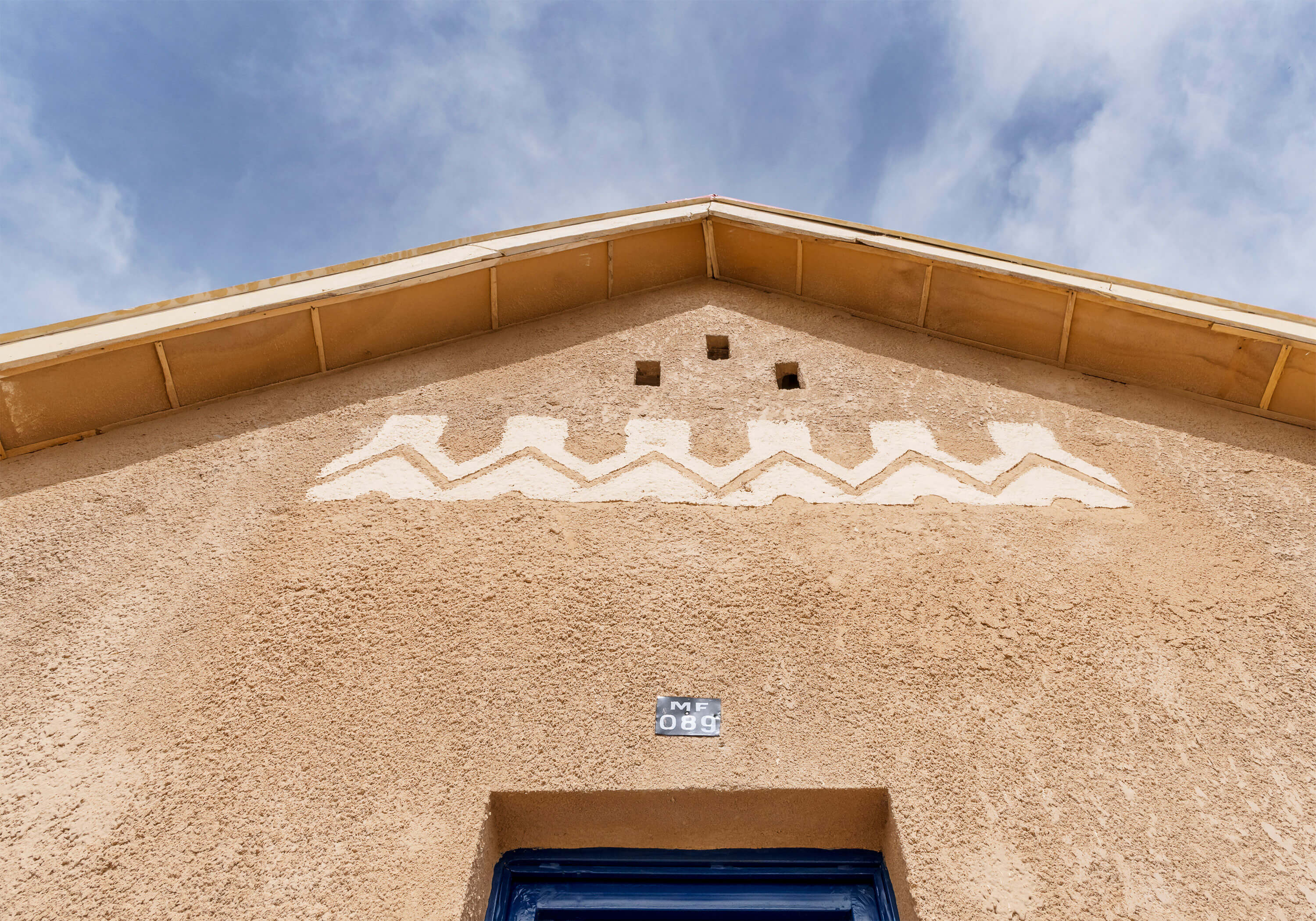 Close-up view of the exterior facade of a building designed by Oshinowo Studio, a leading architectural design studio in Lagos, led by Tosin Oshinowo. The wall is textured with ochre render and adorned with a white geometric frieze under the eaves. Above the striking blue door, three small square windows are set high, allowing light and air while maintaining privacy. The architectural details, including the green metal roof against the blue sky, reflect a blend of local style and modern functionality.