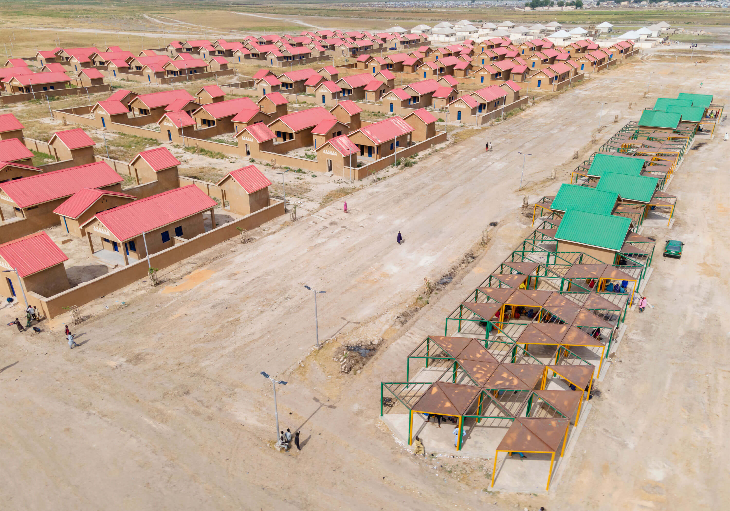Aerial view of a newly developed housing community with designs by Oshinowo Studio, a leading architectural design studio in Lagos, led by Tosin Oshinowo. The area features rows of identical houses with ochre walls and red roofs, organized in a neat grid pattern. A market street with green and yellow metallic shades offers a communal space for trade and socializing. The environment appears arid, with little vegetation, highlighting the development as an oasis of human habitation.