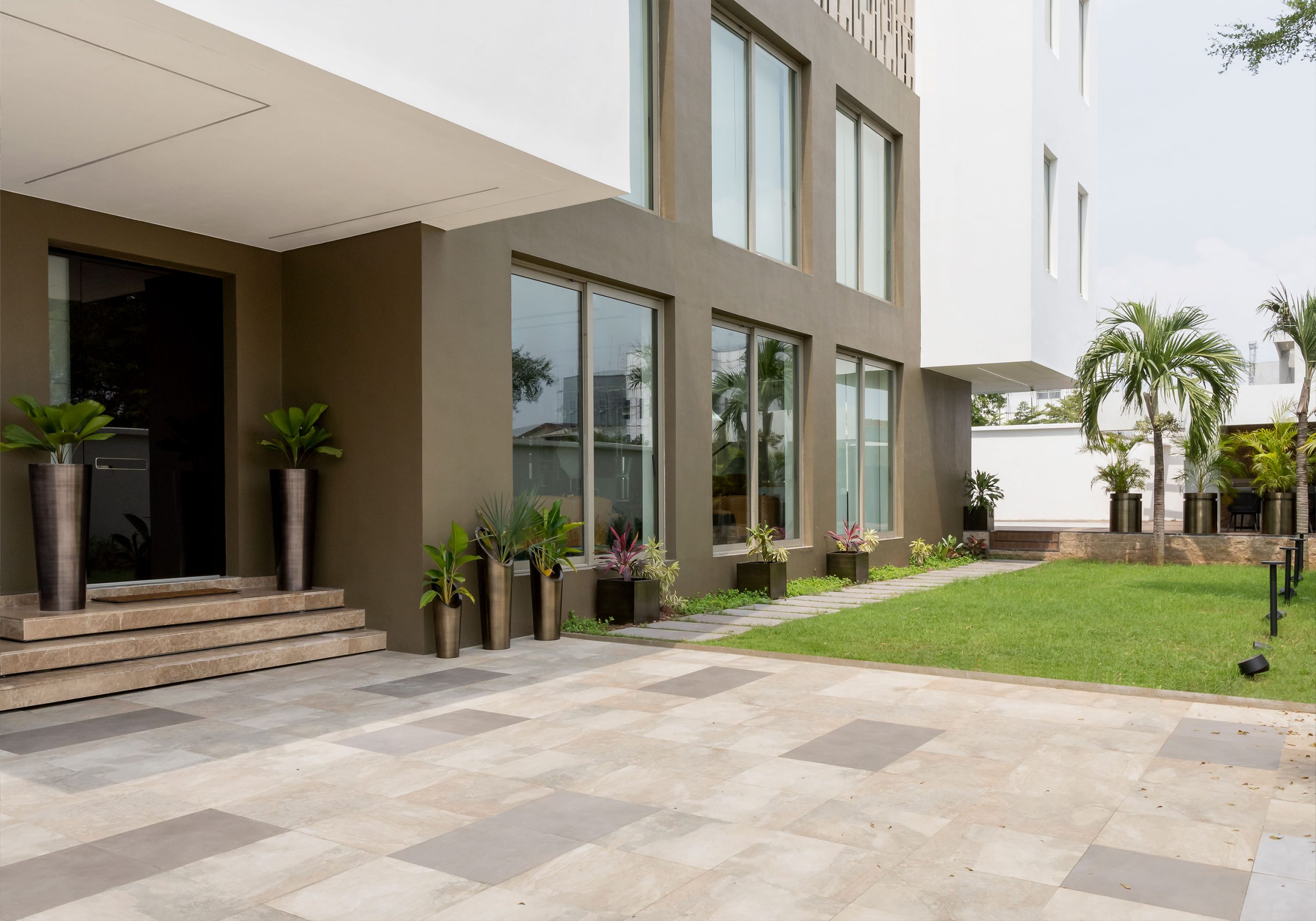 An exterior view of the 'Lantern House' by the leading architectural design studio in Lagos, Oshinowo Studio, led by Tosin Oshinowo, featuring a contemporary design with large windows, a patterned pavement courtyard, and landscaped greenery against the building's modern gray and white facade.