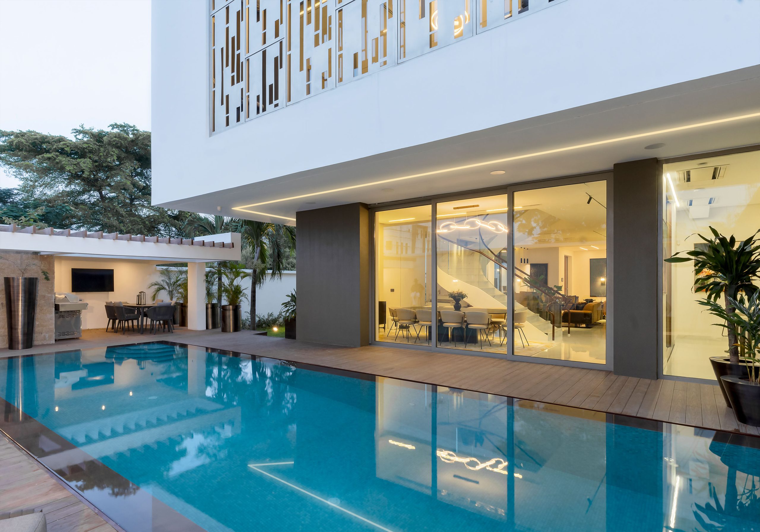 Dusk setting over the 'Lantern House' poolside, designed by the leading architectural design studio in Lagos, Oshinowo Studio, led by Tosin Oshinowo, showcasing the building's elegant white exterior with a decorative metal screen, complemented by soft ambient lighting and a serene, reflective blue pool.