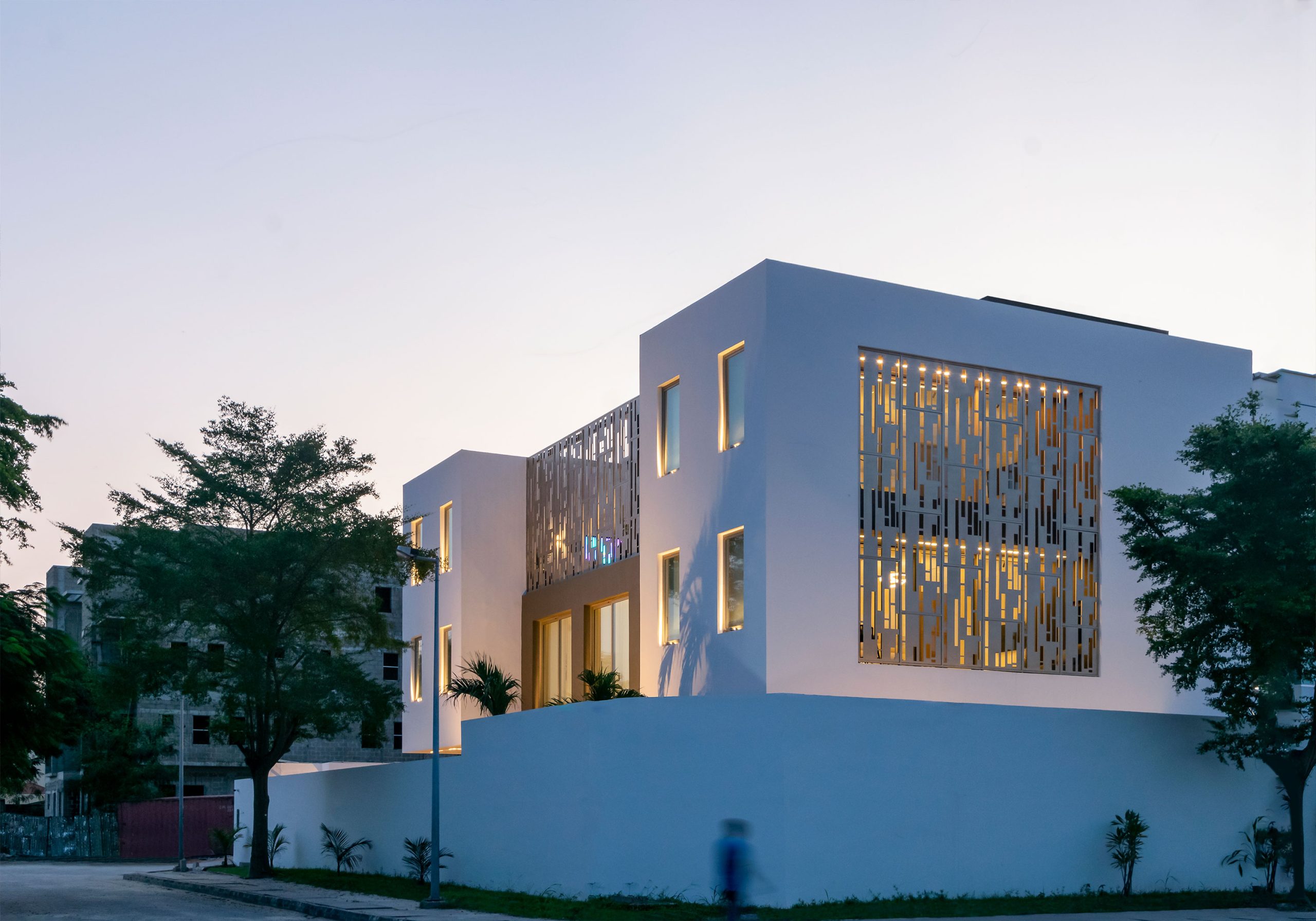 Twilight view of the Lantern House, a contemporary masterpiece by the leading architectural design studio in Lagos, Oshinowo Studio, led by Tosin Oshinowo, with its striking white facade punctuated by elegant golden lighting through the artistic metal screen, nestled amongst the greenery of a tranquil neighborhood.