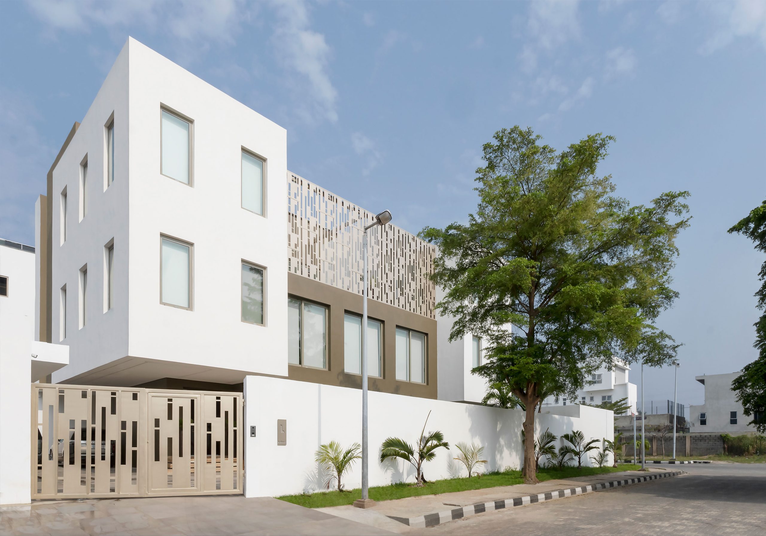 The 'Lantern House' in daylight, designed by the leading architectural design studio in Lagos, Oshinowo Studio, led by Tosin Oshinowo, displaying a striking white facade punctuated by symmetrical windows and an artistic metallic screen, all set against a backdrop of blue sky and lush greenery.