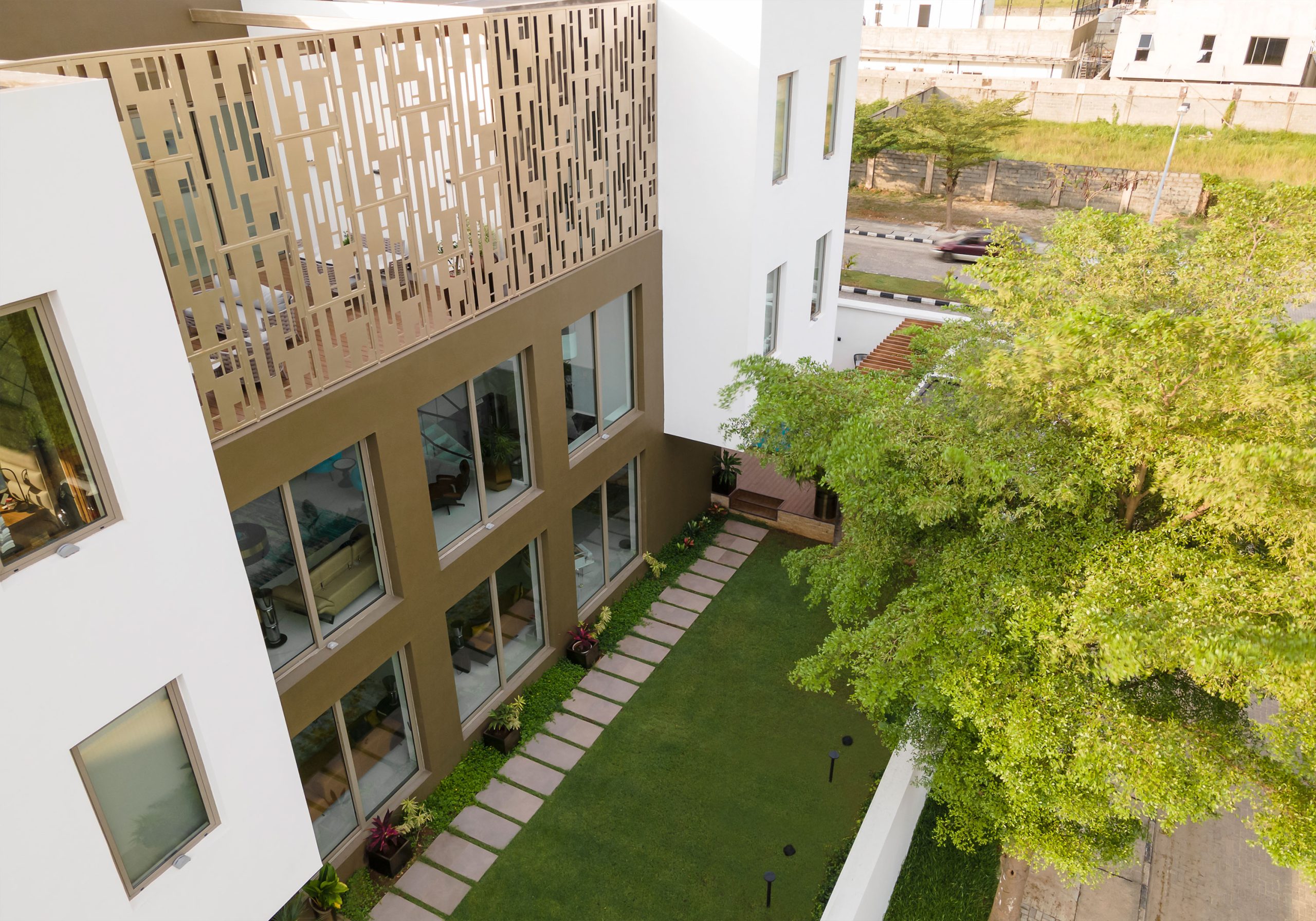 A bird's-eye view of a landscaped courtyard within a residential complex by the leading architectural design studio in Lagos, Oshinowo Studio, led by Tosin Oshinowo, with a beige and white building featuring a unique metallic facade, large windows, and surrounded by lush trees and manicured grass.
