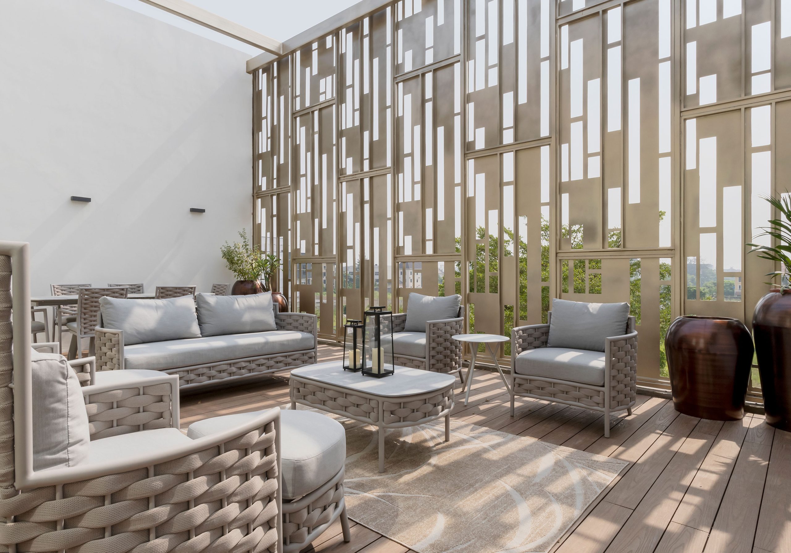 An elegantly furnished outdoor patio area of a property designed by the leading architectural design studio in Lagos, Oshinowo Studio, led by Tosin Oshinowo, with modern wicker furniture, cozy gray cushions, a decorative metal screen providing a dynamic backdrop, and a warm wooden deck underfoot.