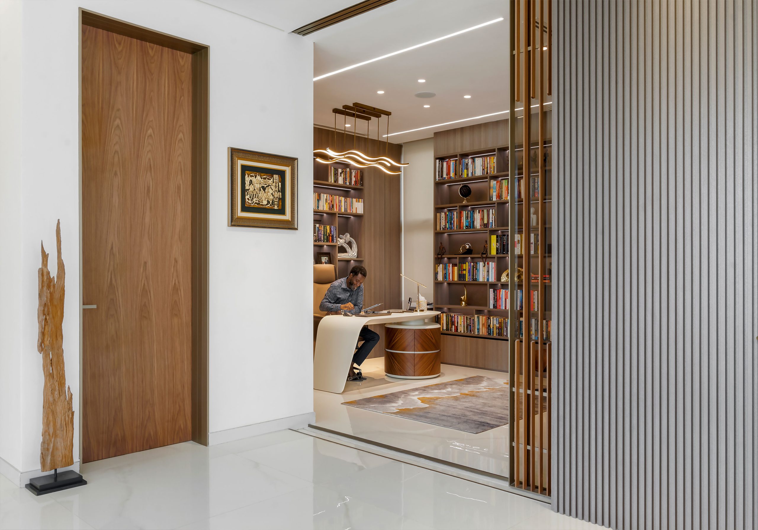 An elegant home office space designed by the leading architectural design studio in Lagos, Oshinowo Studio, featuring a wooden desk, a bookshelf filled with books, unique lighting fixtures, and a person focused on work, with a polished marble floor and decorative pieces that add a touch of sophistication. This project was led by Tosin Oshinowo.