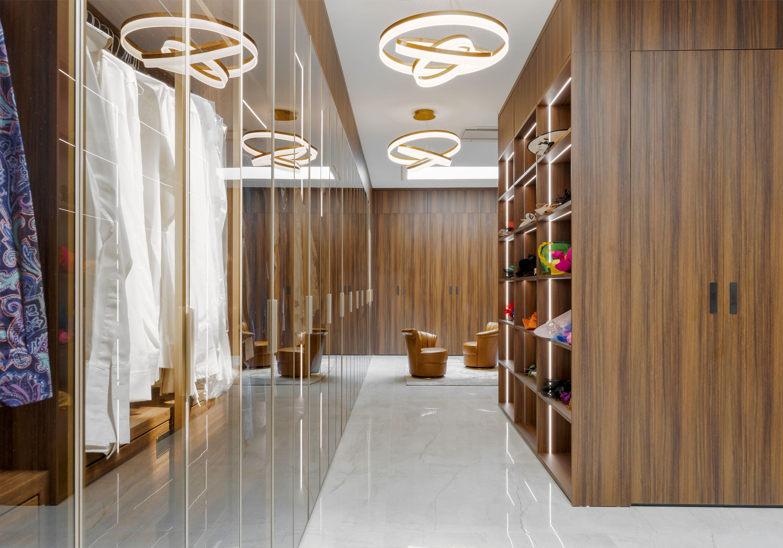 A luxurious and modern walk-in closet design by the leading architectural design studio in Lagos, Oshinowo Studio, led by Tosin Oshinowo, with sleek wood paneling, contemporary circular light fixtures, a mirrored wardrobe, and open shelving displaying a colorful array of items, all over a polished marble floor.