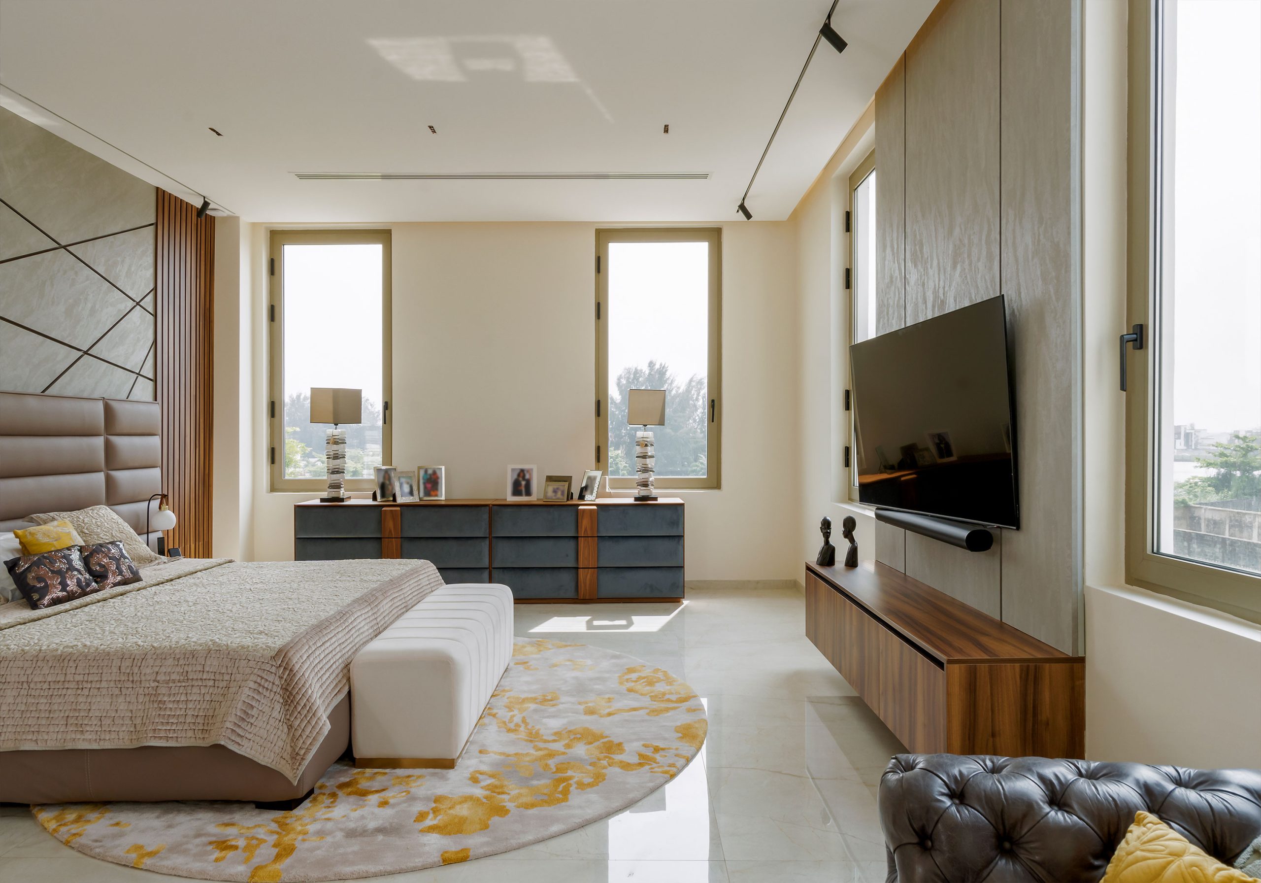 A spacious and well-lit bedroom designed by the leading architectural design studio in Lagos, Oshinowo Studio, led by Tosin Oshinowo, featuring a large bed with a tufted headboard, an elegant round rug with gold accents, a sleek TV console, and a plush leather bench at the foot of the bed, complemented by expansive windows and a harmonious color palette.