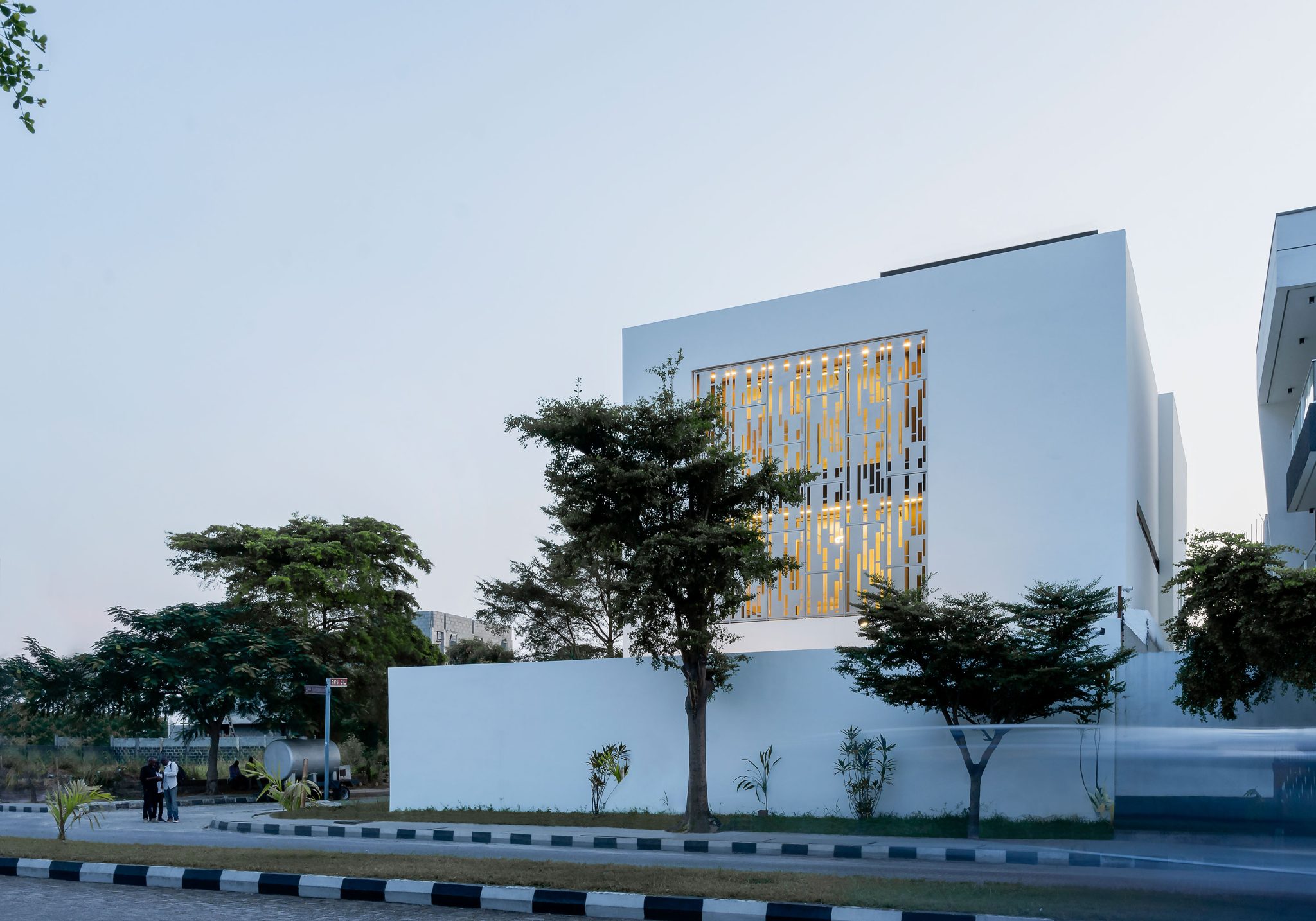 An early evening view of the 'Lantern House' by the leading architectural design studio in Lagos, Oshinowo Studio, led by Tosin Oshinowo, featuring a modern white building with an intricate golden metal screen facade, surrounded by lush green trees and a clear blue sky.