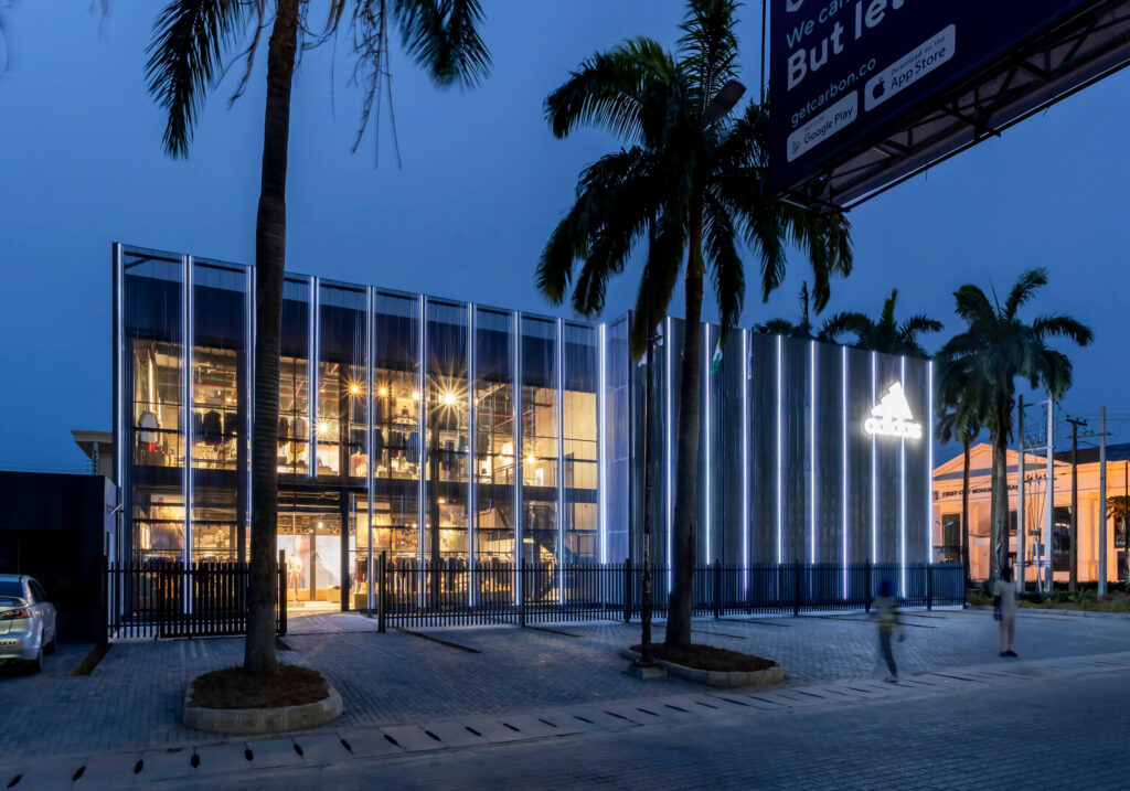 Evening view of the ADIDAS Lagos flagship store, a contemporary two-story building with a transparent glass facade that allows a peek into the brightly lit interior showcasing sportswear and accessories. The store's design, by the leading architectural design studio in Lagos, Oshinowo Studio led by Tosin Oshinowo, features vertical illuminated stripes on the exterior walls creating a dynamic visual effect. Lush palm trees frame the structure, and a prominent ADIDAS logo glows above, casting a soft light on the surroundings. Pedestrians can be seen in motion, emphasizing the store's integration into the city's vibrant street life.